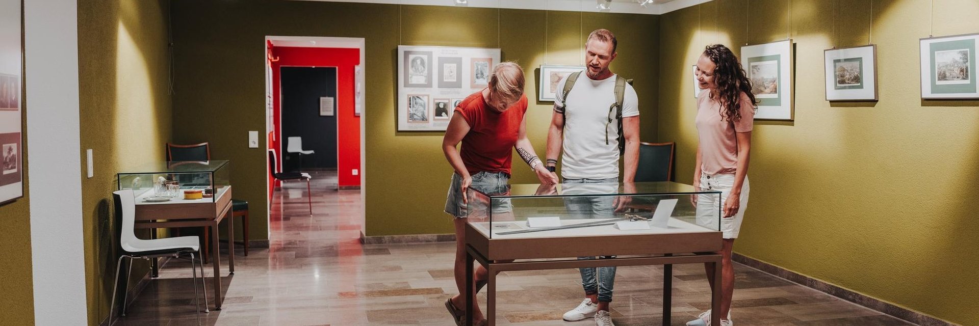 Drei Personen betrachten eine Ausstellungsvitrine in einem Museum mit Bildern an den Wänden und einem roten Türrahmen im Hintergrund