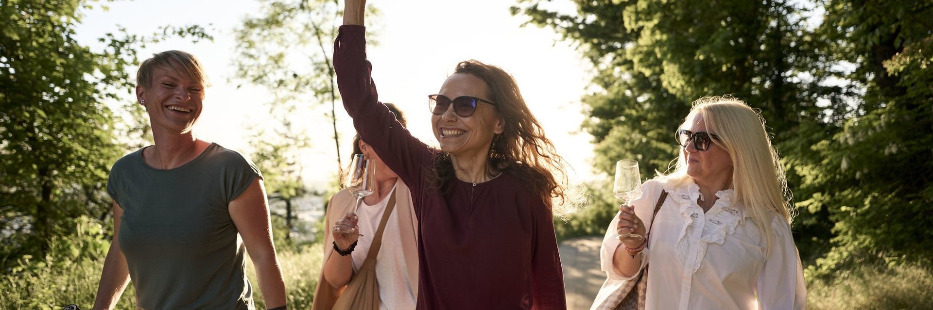 Vier Frauen gehen lachend auf einem Weg, eine hält eine Flasche hoch, alle tragen Sonnenbrillen und Freizeitkleidung