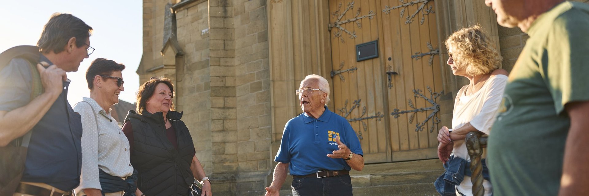 Älterer Mann in blauem Poloshirt spricht vor einer Gruppe von fünf Personen vor einem historischen Gebäude mit steinerner Fassade und großen Holztüren