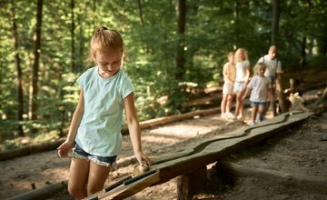 Mädchen mit hellem T-Shirt und kurzen Hosen spielt an einer Murmelbahn im Wald, im Hintergrund stehen mehrere Personen.