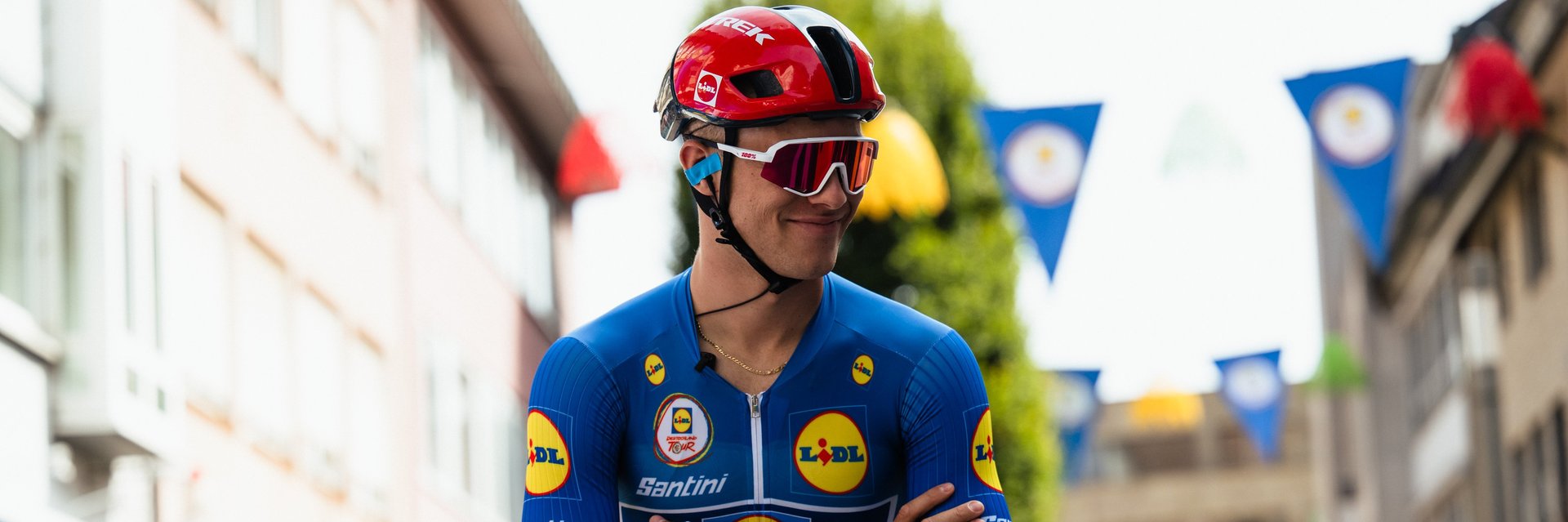 Radrennfahrer in blauem Trikot mit roten Helm und Sonnenbrille sitzt auf Rennrad vor Zuschauern in einer Stadt