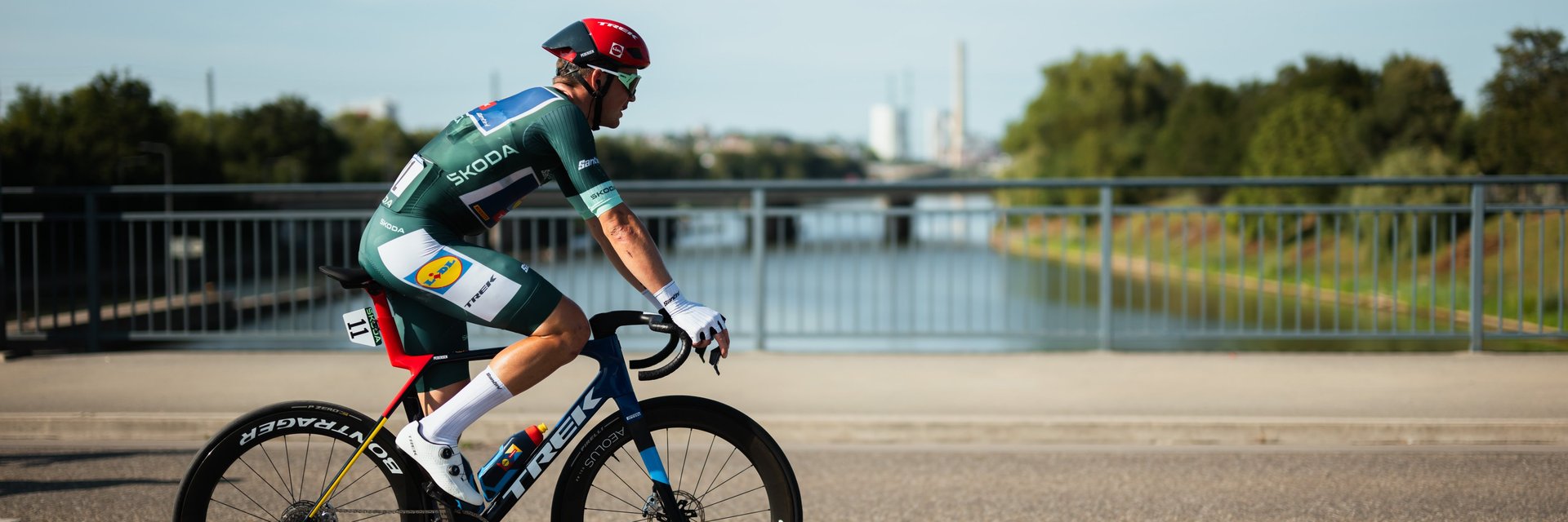 Radrennfahrer in grünem Trikot und rotem Helm fährt auf Rennrad auf einer Brücke mit Fluss im Hintergrund
