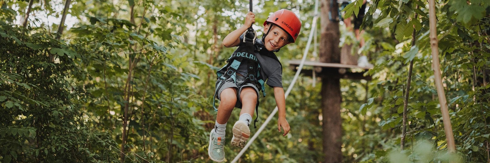 Kind mit rotem Helm und Klettergurt fährt an einer Seilrutsche durch einen bewaldeten Kletterpark