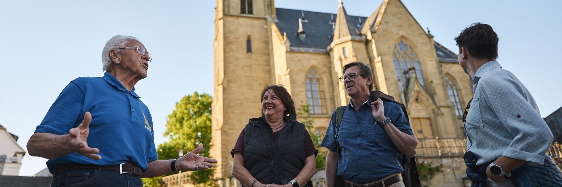 Vier Personen stehen vor einer Kirche und unterhalten sich, ein älterer Mann gestikuliert dabei lebhaft