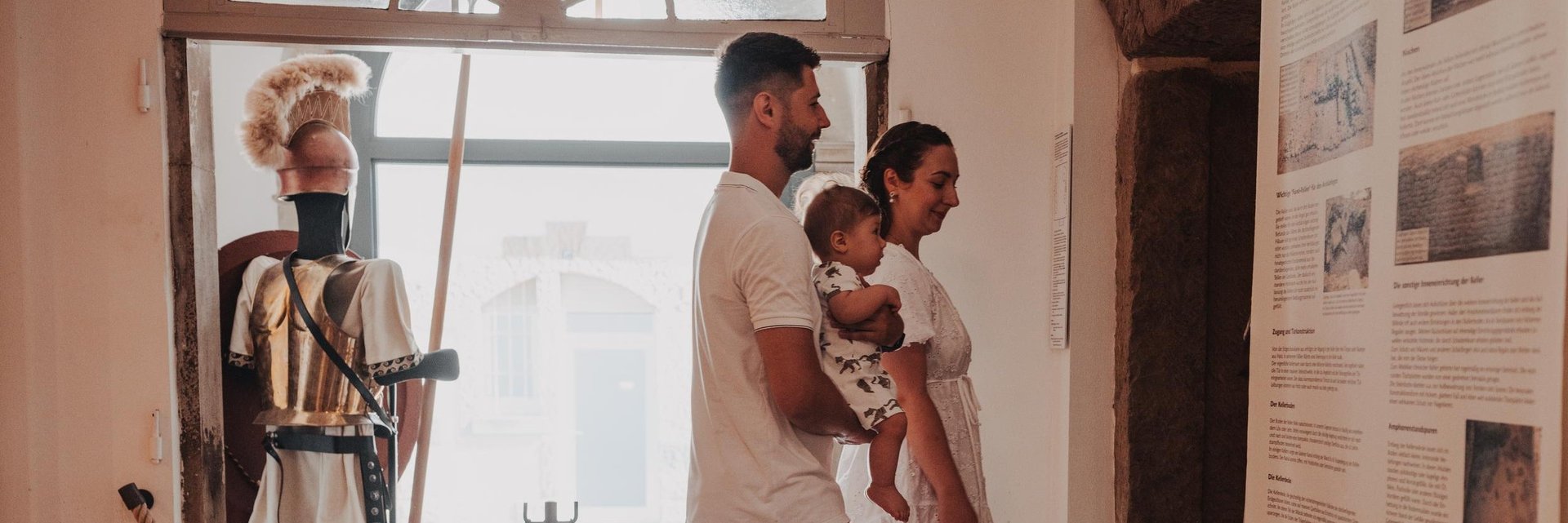 Familie mit Kind betrachtet Ausstellungstafeln in einem Museum mit römischer Rüstung im Hintergrund