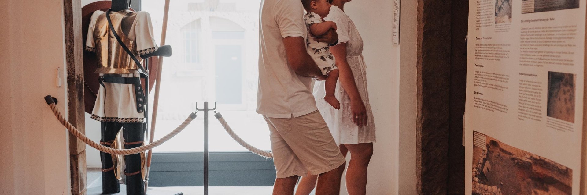 Familie mit Kind betrachtet Ausstellungstafeln in einem Museum mit römischer Rüstung im Hintergrund