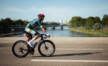 Radrennfahrer in grünem Trikot und rotem Helm fährt auf Rennrad auf einer Brücke über einen Fluss