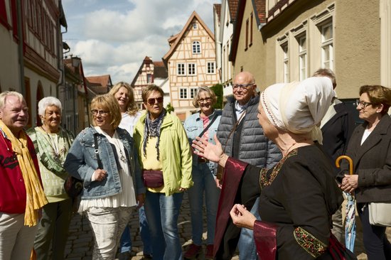 Frau in historischer Tracht mit weißer Haube spricht vor einer Gruppe von Menschen auf einer gepflasterten Straße in einer Altstadt