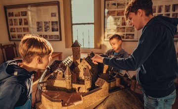 Drei Jungen betrachten und berühren ein detailliertes Modell einer Burg in einem Raum mit Fenstern und gerahmten Bildern an den Wänden