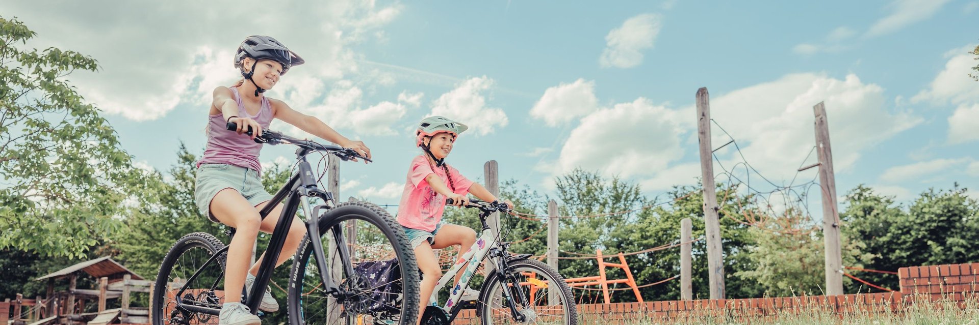 Imagebild Familien-Radtouren | Zwei Mädchen auf dem Fahrrad fahren an einem Spielplatz vorbei.
