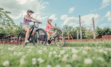 Imagebild Familien-Radtouren | Zwei Mädchen auf dem Fahrrad fahren an einem Spielplatz vorbei.