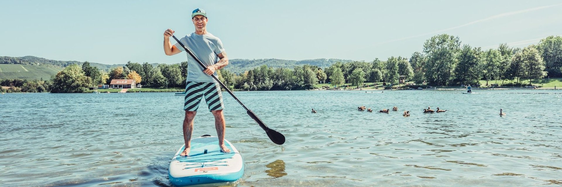 Mann steht auf einem Stand-up-Paddle-Board im flachen Wasser eines Sees und hält ein Paddel