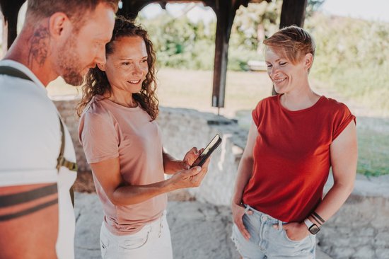Drei Personen stehen unter einem Holzdach, eine Frau zeigt den anderen etwas auf ihrem Smartphone