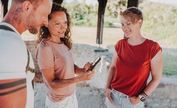 Drei Personen stehen unter einem Holzdach, eine Frau zeigt den anderen etwas auf ihrem Smartphone