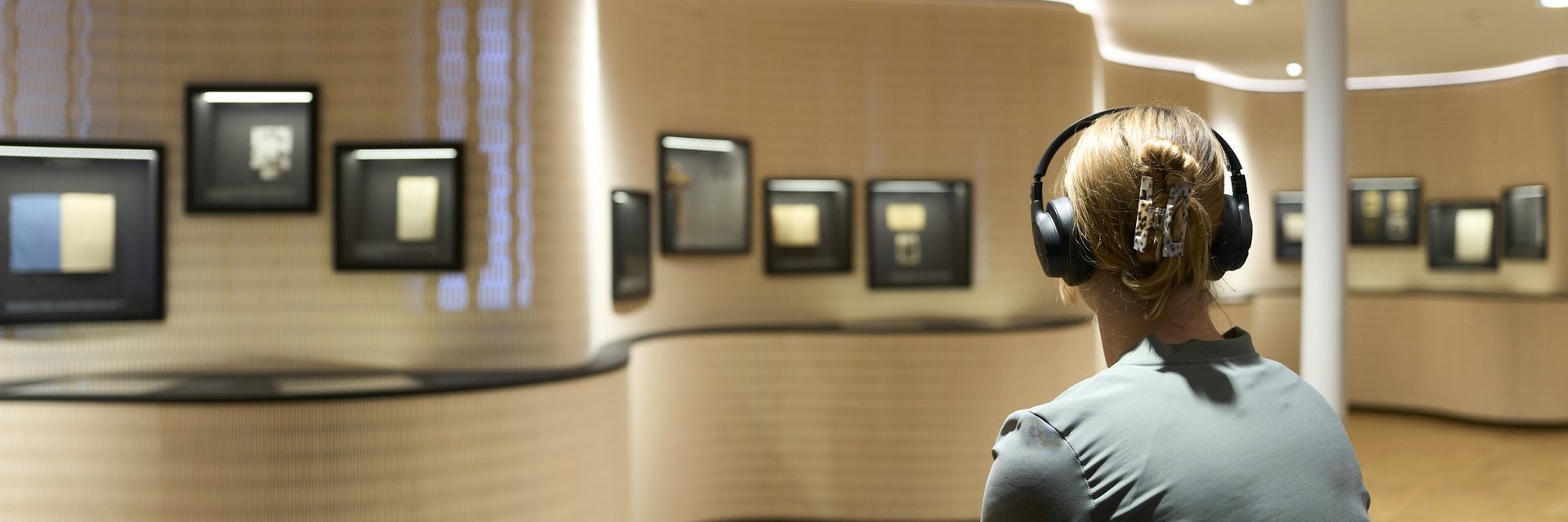 Person mit Kopfhörern sitzt in einem Museum vor einer geschwungenen Wand mit mehreren gerahmten Bildern
