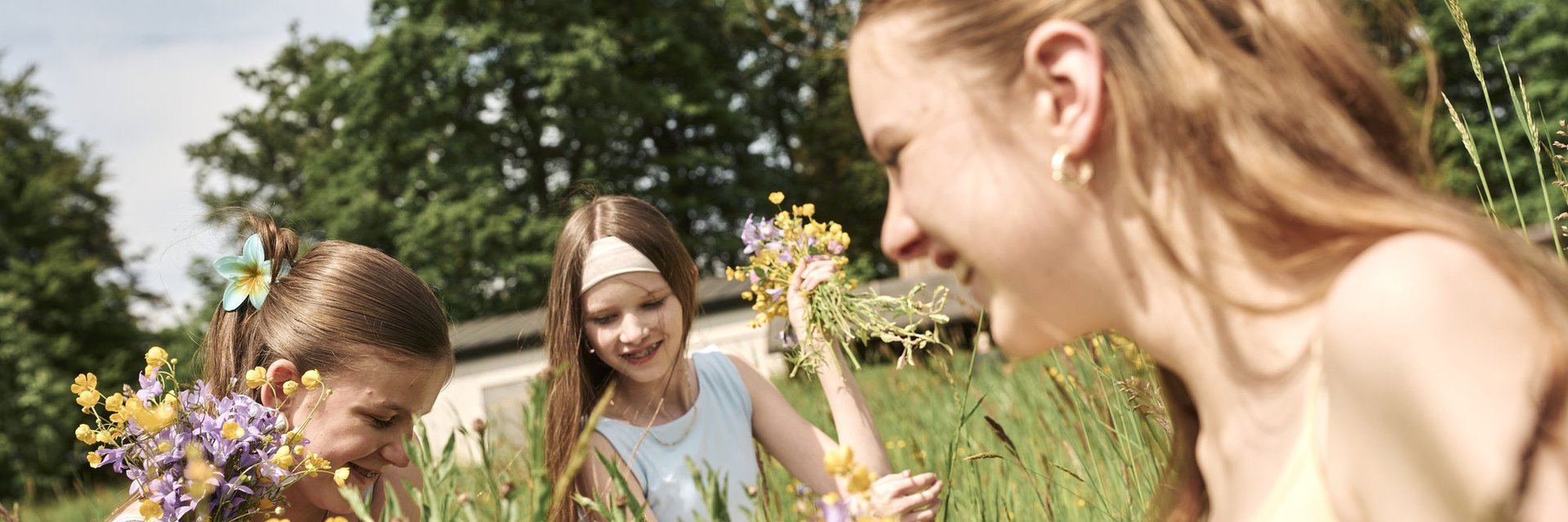 Drei Mädchen mit Blumensträußen sitzend lachend auf einer Wiese vor einem hölzernen Aussichtsturm
